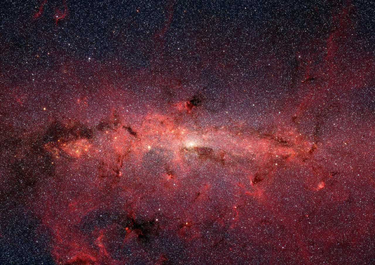 Middle third of Spitzer Space Telescope infrared image shows a horizontal strip of stars and reddish, glowing gas and tendrils of dark dust.